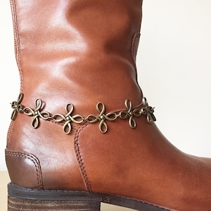 Könnte beinhalten: Ein brauner Lederstiefel mit einer goldenen Kette und dekorativen Metallanhängern um den Knöchel.