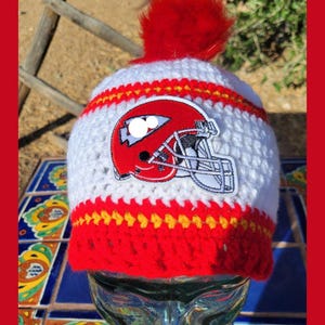 May include: A handmade, crocheted winter hat in white, red, and yellow. The hat features a red pom-pom on top and a patch of a red football helmet. The hat is designed with alternating white and red stripes.