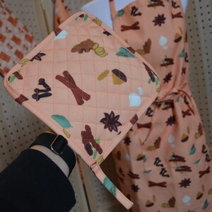 May include: Peach-colored quilted pot holder with a loop, featuring a pattern of brown cinnamon sticks, star anise, and other spice illustrations. An apron with the same pattern is visible in the background. The pot holder is square-shaped.