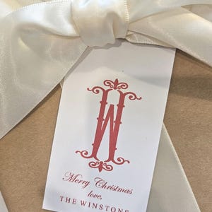 May include: A white gift tag with a red monogram letter "W" and the words "Merry Christmas love, THE WINSTONS". The tag is tied with a cream-colored satin ribbon, creating a festive holiday presentation.