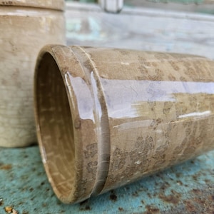 May include: Two antique, beige ceramic pots with a glossy finish. One pot is upright, the other lying on its side. The pots have a textured surface and a cylindrical shape. They are likely for decorative or functional use, possibly for plants.