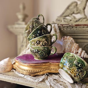 Può includere: Una pila di cinque tazze da tè in ceramica verde e bianca con motivi floreali e conchiglie. Le tazze sono impilate su un vassoio di velluto viola e dorato.