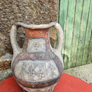 Peut inclure: Un vase en céramique à deux anses, avec un corps large et un col étroit. Le vase présente un motif géométrique en noir, blanc et marron. Le vase est posé sur une surface rouge, avec un mur en pierre et une porte en bois verte en arrière-plan.