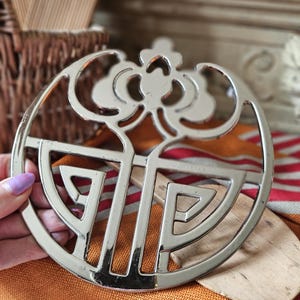 May include: A silver metal trivet with a decorative floral design. The trivet is round and has a geometric pattern.