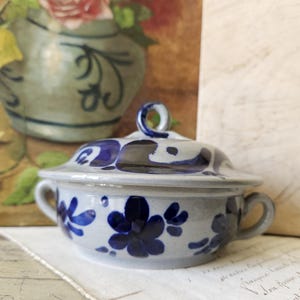 May include: A light gray ceramic lidded bowl featuring hand-painted dark blue floral patterns. The bowl has two small side handles and a matching lid with a circular loop handle on top. The glaze has a speckled texture throughout the surface.