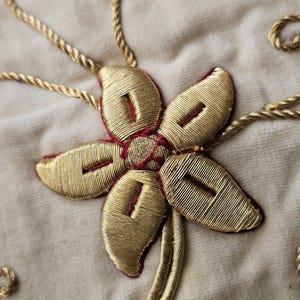 May include: Close-up of a gold embroidered flower with five petals and a red center. The flower is stitched onto a cream-colored fabric. Gold rope-like embroidery surrounds the flower, with additional decorative swirls.