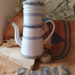 Könnte beinhalten: Eine weiße Emaille-Kaffeekanne mit blauen und goldenen Streifen. Die Kanne hat einen Deckel und einen Ausguss. Sie steht auf einem Holzbrett.