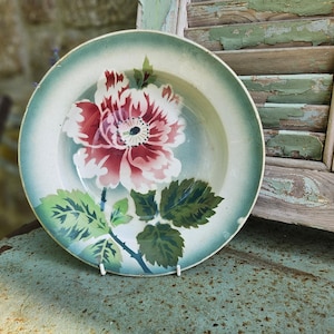 Puede incluir: Un plato de cerámica vintage con un diseño floral. El plato presenta una gran flor roja y blanca con hojas verdes sobre un fondo azul claro. El plato se exhibe sobre una superficie metálica.