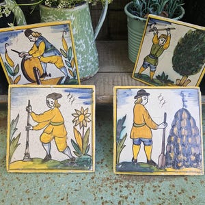 Puede incluir: Cuatro baldosas de cerámica cuadradas con ilustraciones pintadas a mano de figuras con ropa amarilla y azul. Cada baldosa representa una escena diferente, incluyendo una persona afilando una herramienta, cortando un árbol y trabajando con un pajar.