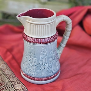 May include: A vintage ceramic pitcher with a cream-colored top and handle, a light blue body, and a burgundy rim and interior. The body features a relief scene of figures. The handle has a textured, braided design. The pitcher is displayed on a red cloth.