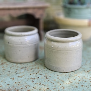 Puede incluir: Dos vasijas cilíndricas de gres con un esmalte de color beige claro moteado. Cada recipiente presenta un borde liso y redondeado, junto con una sutil hendidura horizontal que rodea la parte superior del cuerpo, mostrando un diseño funcional.
