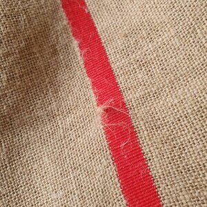 May include: Close-up of a natural burlap fabric with a single red stripe running diagonally across the surface.