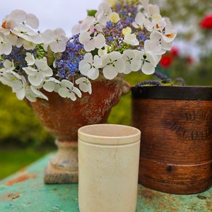 May include: A white ceramic cup with a rustic brown and white planter filled with white and purple flowers. The planter is sitting on a green wooden table.