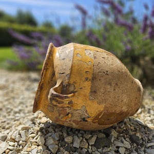 Puede incluir: Una maceta grande de terracota amarilla con un acabado rústico desgastado. La maceta está tumbada sobre un lecho de pequeñas piedras grises.