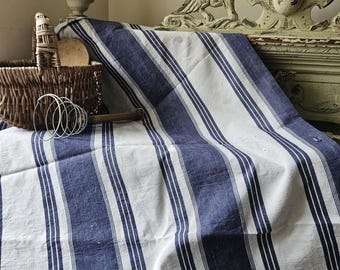 Peut inclure: Une nappe en lin à rayures bleues et blanches. Un panier en osier, une cuillère en bois et un fouet métallique sont au premier plan. L'arrière-plan présente une tête de lit décorative en bois blanc cassé vieilli.