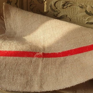 May include: A piece of natural burlap fabric with a single red stripe running across the width. The fabric is folded and has a rough, textured surface.