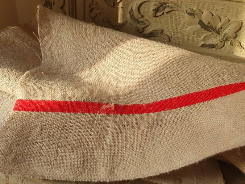 May include: A piece of natural burlap fabric with a single red stripe running across the width. The fabric is folded and has a rough, textured surface.