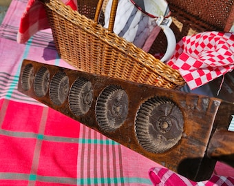 Op de afbeelding: Een houten mal met zes ronde indrukken, elk met een gedetailleerd ontwerp. De mal is bruin en heeft een natuurlijke houten afwerking. Hij ligt op een rood-wit geblokt tafelkleed.