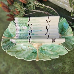 May include: A ceramic asparagus serving dish. The dish is shaped like a large leaf with a raised section designed to hold asparagus spears. The asparagus is painted in shades of green and purple, with a cream-coloured base. The leaf-shaped dish is green.