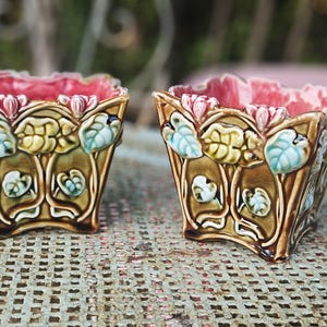 May include: Two square ceramic planters featuring a relief design of raised leaves and flowers. The exterior is glazed in shades of brown, light blue, and yellow, while the interior is finished in a vibrant pink glaze with a scalloped rim.