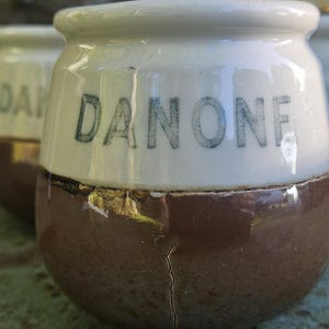 May include: Close-up of three vintage Danone yogurt jars. The jars are white on top and brown on the bottom, with the word "DANONE" printed in green. The jars have a rounded shape and a crack on one of them.