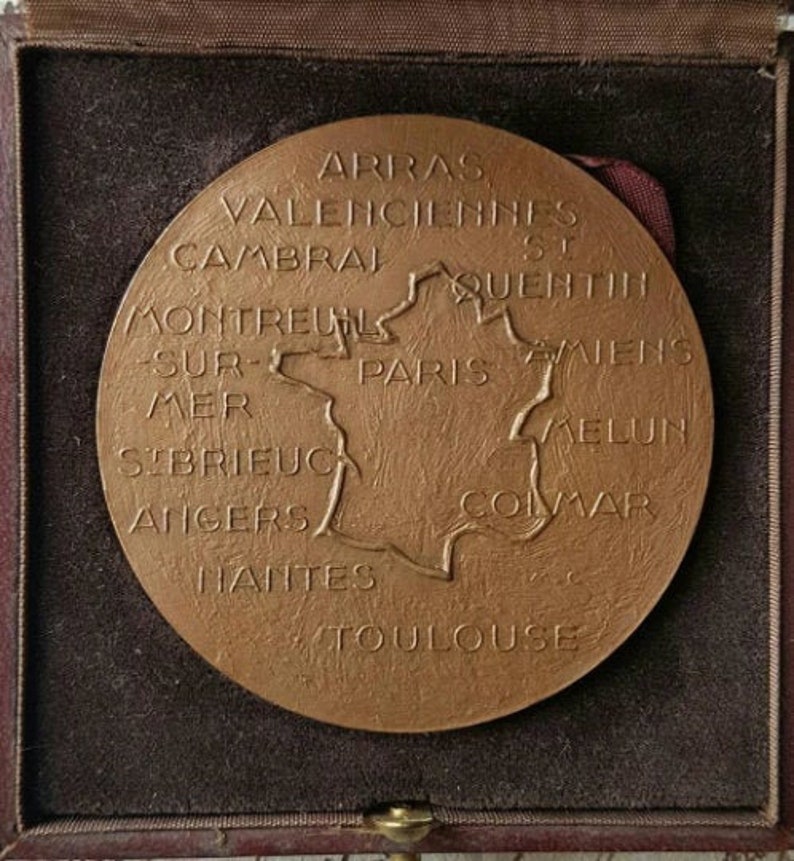 Puede incluir: Una medalla de bronce con un mapa de Francia y los nombres de las ciudades alrededor del per&iacute;metro. Las ciudades que aparecen son: Arras, Valenciennes, Cambrai, St Quentin, Montreuil-sur-Mer, Par&iacute;s, Amiens, Melun, Colmar, Angers, Nantes y Toulouse.