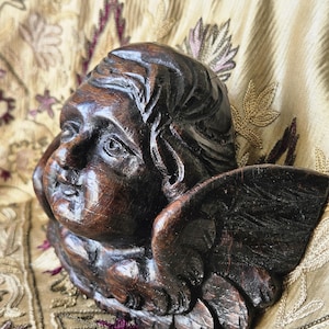 Könnte beinhalten: Eine geschnitzte Holzskulptur eines Engelskopfs mit detaillierten Gesichtszügen und Flügeln. Die Skulptur ist dunkelbraun und scheint antik zu sein. Der Hintergrund ist ein gemusterter Stoff mit gold- und lilafarbenen Stickereien.