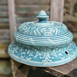 May include: A turquoise ceramic decorative jar with a lid. The jar features an ornate, embossed floral design and a small hole. It sits on a wooden surface. The jar's dimensions are approximately 15 cm in diameter.