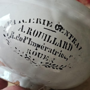 Peut inclure: Gros plan sur un bol en céramique blanc avec texte noir. Le texte comprend le nom "A. ROUILLARD" et les mots "R. de l'Impératrice, ROUEN." Le bol a un bord légèrement ébréché et un aspect rustique.