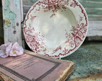 May include: A vintage, off-white ceramic bowl with a red bird and floral pattern. The bowl is placed next to a weathered book and pink flowers. The background includes a distressed, light blue shutter.