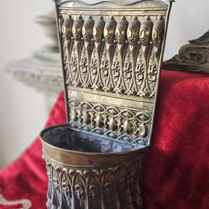 May include: A wall-mounted brass planter featuring an ornate backplate with vertical embossed floral and teardrop motifs. The semi-circular basin has a matching decorative exterior with repeating vertical patterns and a weathered patina finish.