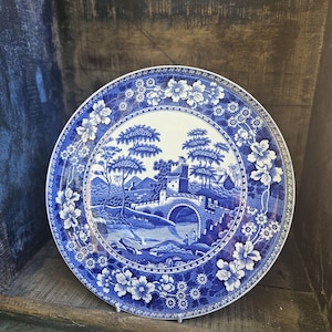 Könnte beinhalten: Blau-weiße Zierteller mit einer Landschaftsszene, die eine Brücke, Bäume und Gebäude zeigt. Der Teller hat einen floralen Rand und wird auf einer Holzoberfläche präsentiert. Wahrscheinlich aus Keramik.