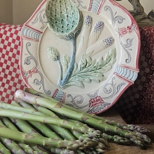 Op de afbeelding: Een decoratieve keramische schaal met een artisjok- en aspergemotief. De schaal heeft een crèmekleurige basis met een roze en blauwe rand. Verse groene asperges liggen op de voorgrond, wat wijst op een culinair thema.
