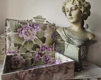 May include: An open vintage floral patterned box with a light green and purple floral design. The box is made of fabric and has a wooden frame. The box is sitting on a white surface.