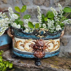 Può includere: Un vaso in ceramica blu e bianca con dettagli dorati decorativi e un viso grottesco al centro. Il vaso è pieno di fiori bianchi e foglie verdi.