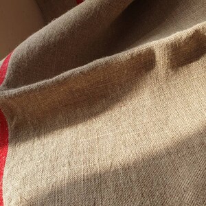 May include: A close-up of a natural linen fabric with a red stripe running down the side. The fabric is textured and has a rustic look.