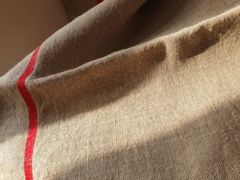 May include: A close-up of a natural linen fabric with a red stripe running down the side. The fabric is textured and has a rustic look.
