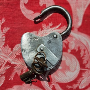 May include: A silver padlock with a heart-shaped body and a key attached. The padlock is on a red and white patterned fabric.