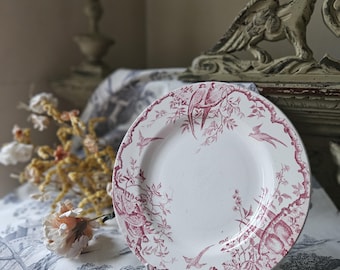 Puede incluir: Un plato de cerámica blanco con un diseño floral y de pájaros rosa. El plato tiene un borde festoneado y está decorado con un patrón repetitivo de pájaros, flores y hojas.
