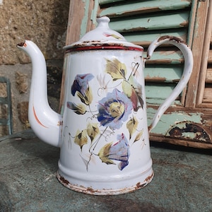 Peut inclure: Cafetière en émail blanc avec un bord rouge et un motif floral. Les fleurs sont bleues, jaunes et vertes. La cafetière a une poignée et un bec.
