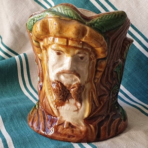 Peut inclure: Pot de fleurs en céramique avec un visage sculpté. Principalement marron, avec des touches de vert et de beige. Le visage porte une barbe et un chapeau, sur un fond de tronc d'arbre.