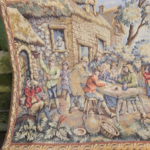 May include: A tapestry depicting a scene of people gathered outside a stone building with a thatched roof. The people are dressed in medieval clothing and are drinking and talking. The tapestry is framed in a brown rope border.