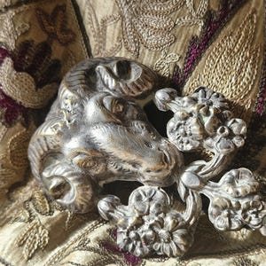 May include: A silver-toned metal ram head sculpture with floral accents. The ram's head is detailed with curled horns and a textured surface. The sculpture is set against a backdrop of embroidered fabric with floral and leaf patterns.