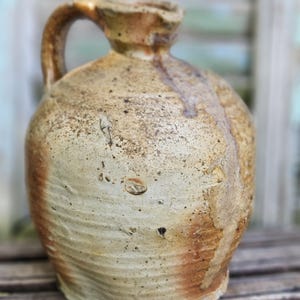 May include: A stoneware jug with a rounded body and a single handle. The surface features a mottled beige and tan glaze with visible speckling, horizontal ridges along the lower section, and a darker brown drip pattern running down the side.