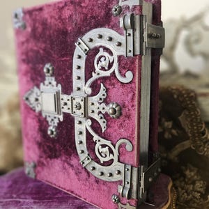 May include: A burgundy velvet-covered book with ornate metal clasps and hinges. The clasps are decorated with intricate floral designs and studded with metal rivets.