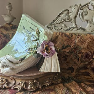 May include: A porcelain bookend shaped like an open book with a pink flower and green leaves. The bookend is on a wooden base with ornate details. The bookend is titled "Oeuvres de Victor Hugo".