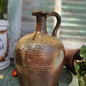 Puede incluir: Una jarra de cerámica rústica hecha a mano con asa. La jarra presenta una combinación de colores marrón y beige con una superficie texturizada y un diseño acanalado. La jarra probablemente se utiliza con fines decorativos.
