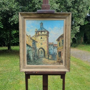 May include: A framed oil painting depicting a European village scene with a tall tower and a clock. The painting is on an easel in front of a green lawn.