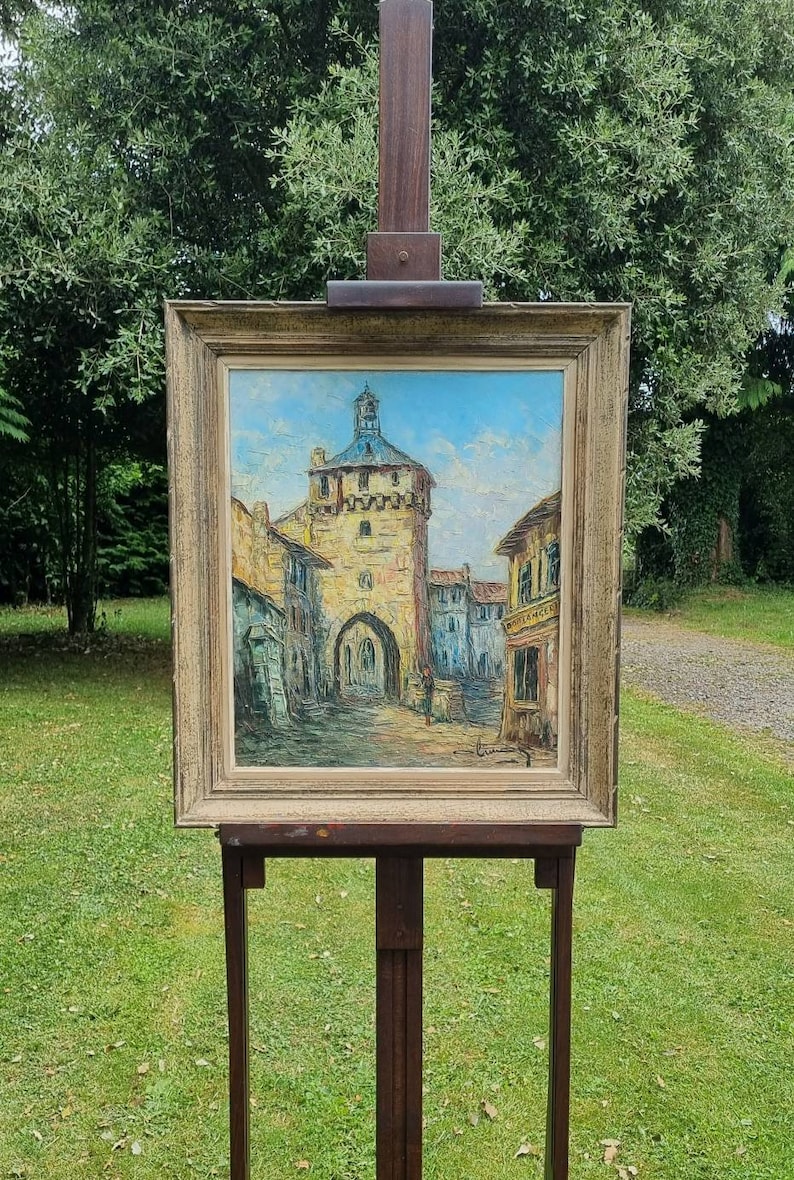 May include: A framed oil painting depicting a European village scene with a tall tower and a clock. The painting is on an easel in front of a green lawn.