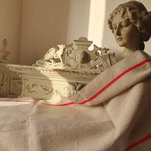 May include: A white, ornate wooden frame with carved details, draped with a beige linen fabric with red stripes. The fabric is folded over the frame and hangs down on both sides.